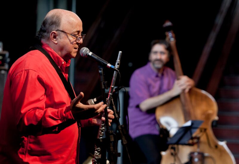 Roberto Sion toca flauta e se apresenta ao microfone, acompanhado por músico que toca contrabaixo acústico.
