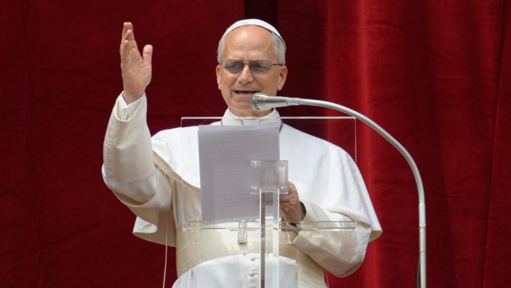 Papa Leão XIV, vestido de branco, acena ao público diante de um microfone, no Vaticano.